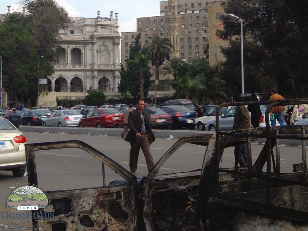 A man walks down the street with a burned out car in the foreground. Cairo